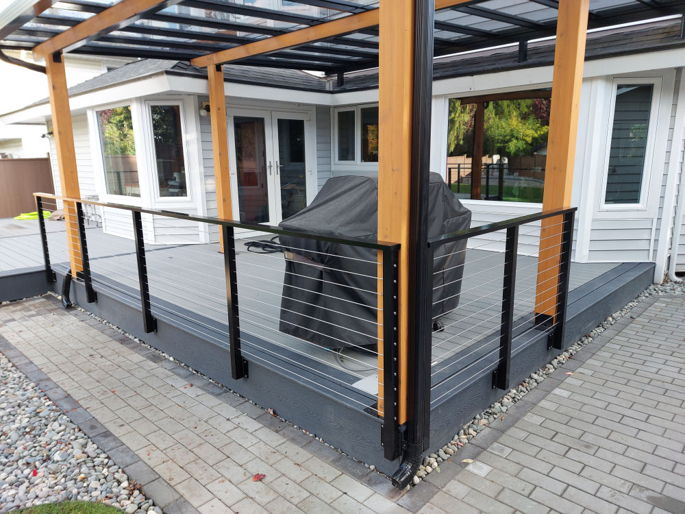 Covered patio with a grill under a cover, framed by wooden posts with a metal and cable railing. The patio floor is gray and the surrounding area is paved with stones.