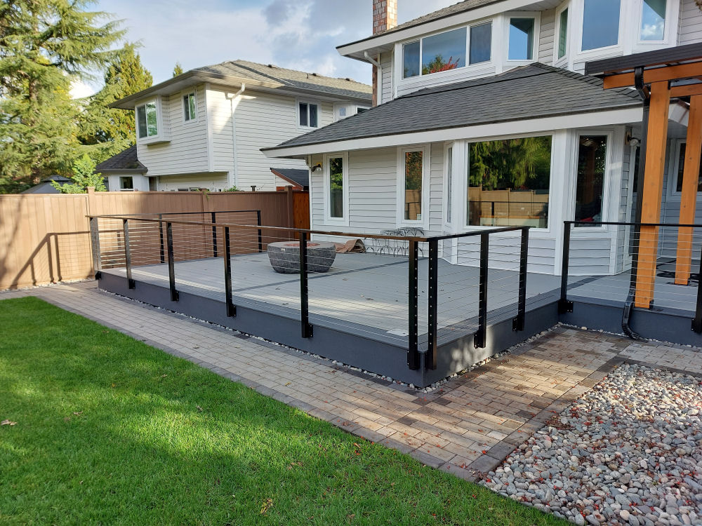 Modern backyard patio with glass railing on a wooden deck attached to a white house. It features a small stone fire pit and is bordered by a brown fence and well-kept lawn.