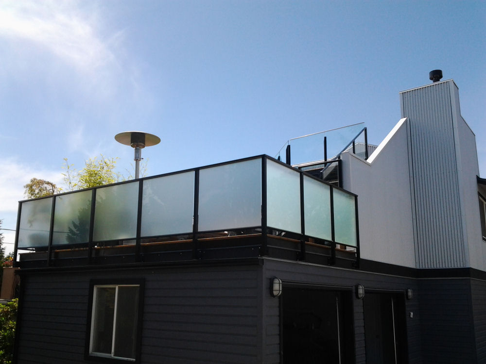 Modern two-story house exterior with a glass-paneled rooftop patio, gray siding, and white chimney against a clear blue sky.