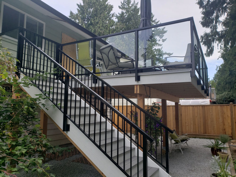 Elevated patio with glass railing and outdoor furniture, accessed by a staircase with black railings. Surrounded by trees and a wooden fence in a backyard setting.