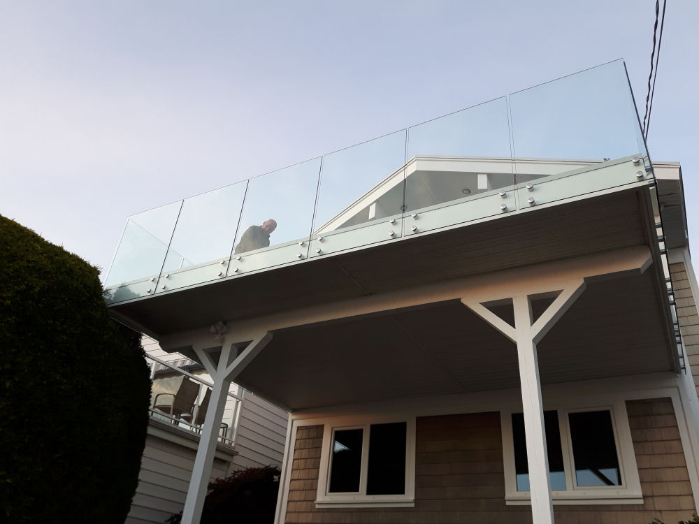House with a second-floor glass balcony, where a person stands looking outward. The structure is supported by white beams, and there's a large bush on the left.