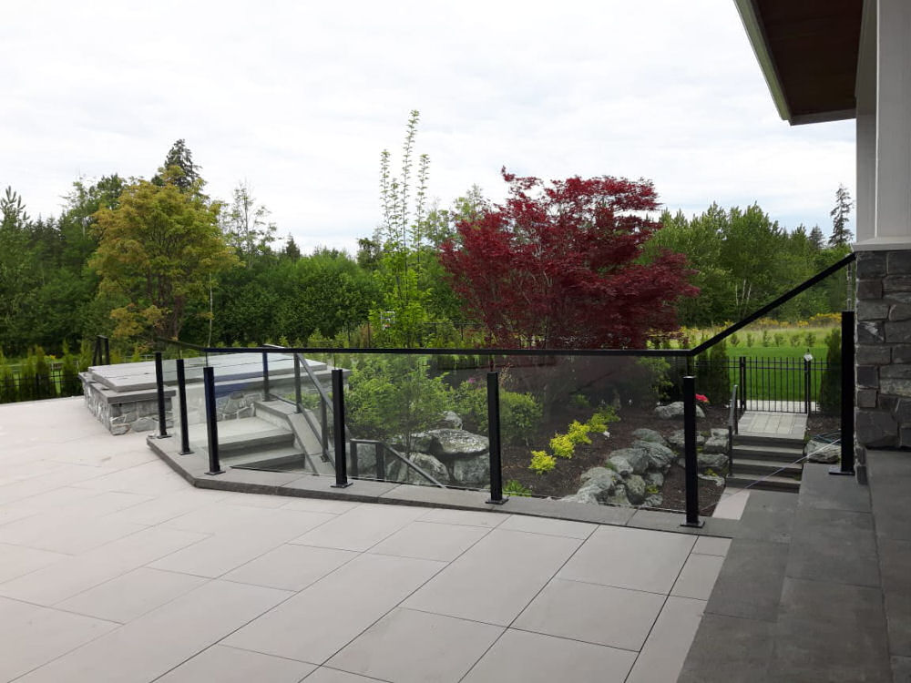 Spacious patio with light tiles and glass railing. Steps lead to a landscaped garden featuring red and green foliage, with trees in the background.