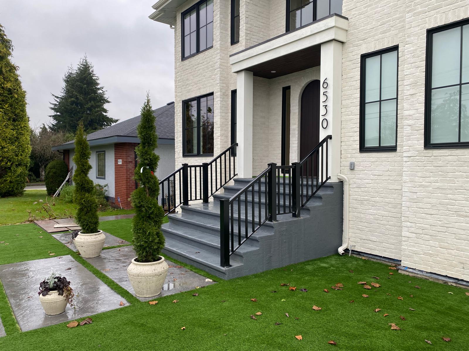 Front view of a modern house with a white brick exterior, black-framed windows, and a covered entrance. Potted plants and artificial grass adorn the front yard, complemented by sleek railings. House number 6530 is displayed.
