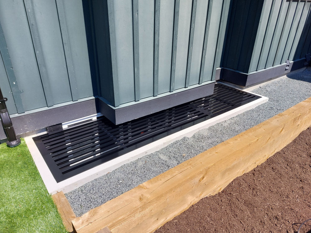 A black metal vent is installed at the base of a blue-paneled building, bordered by concrete and gravel, with a wooden edge leading to a patch of soil.