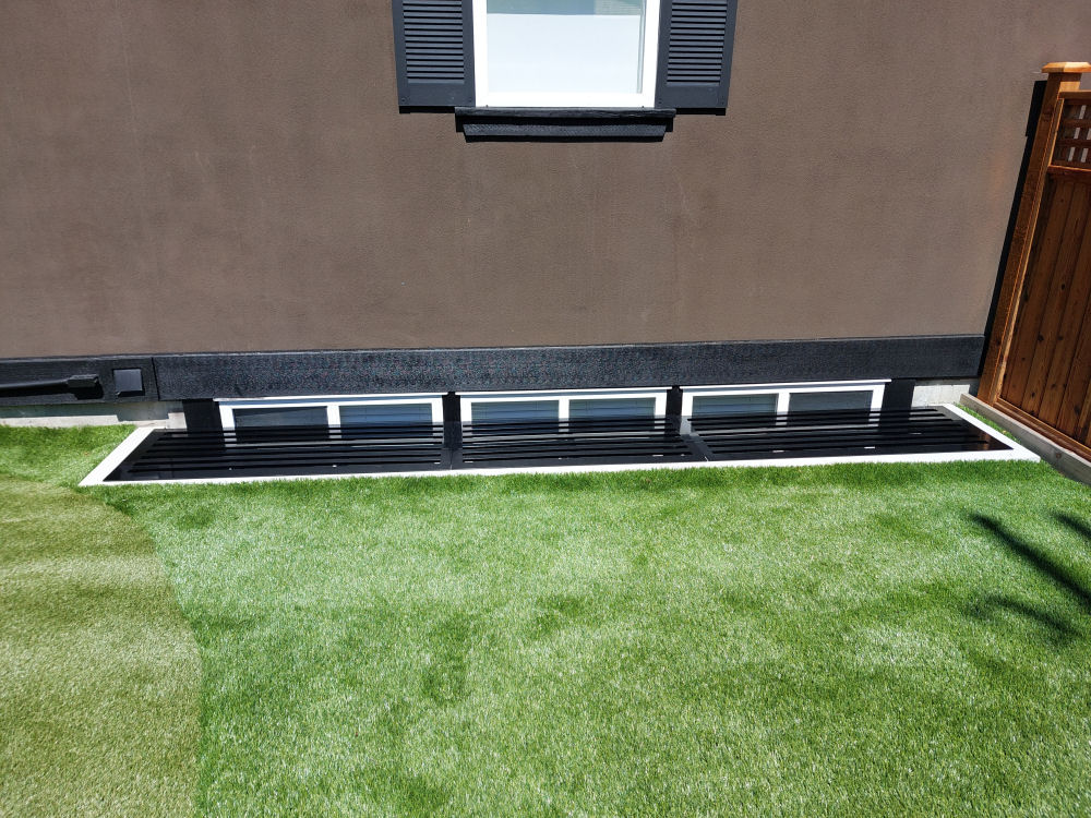 Exterior view of a basement window well covered with a black metal grate, surrounded by artificial grass and next to a wooden fence.