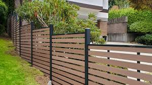 Modern horizontal wooden fence with black vertical posts, bordering a garden with green plants and shrubs.