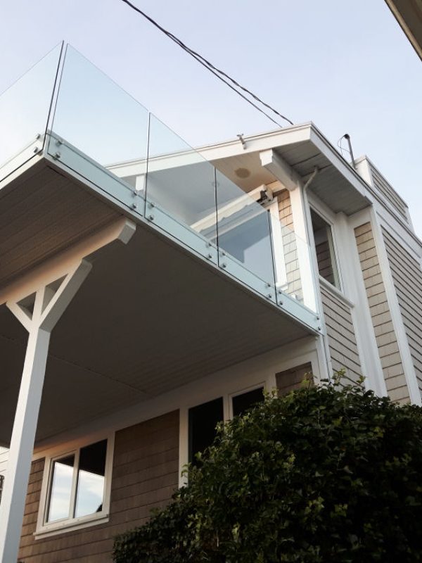 Exterior view of a modern two-story house with a glass balcony and beige siding, taken from a low angle. A tree is partially visible in the foreground.
