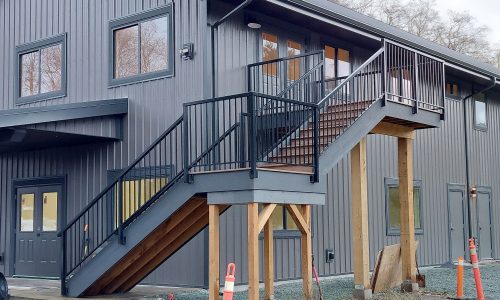 Exterior metal staircase with wooden support beams attached to a gray building. Orange traffic cones are placed nearby on a gravel path.